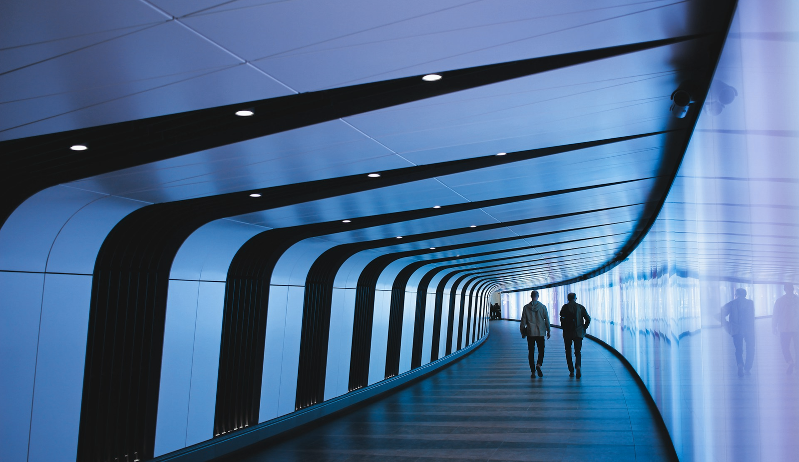 Two men walking down a long, curved hallway lit in blueish purple.